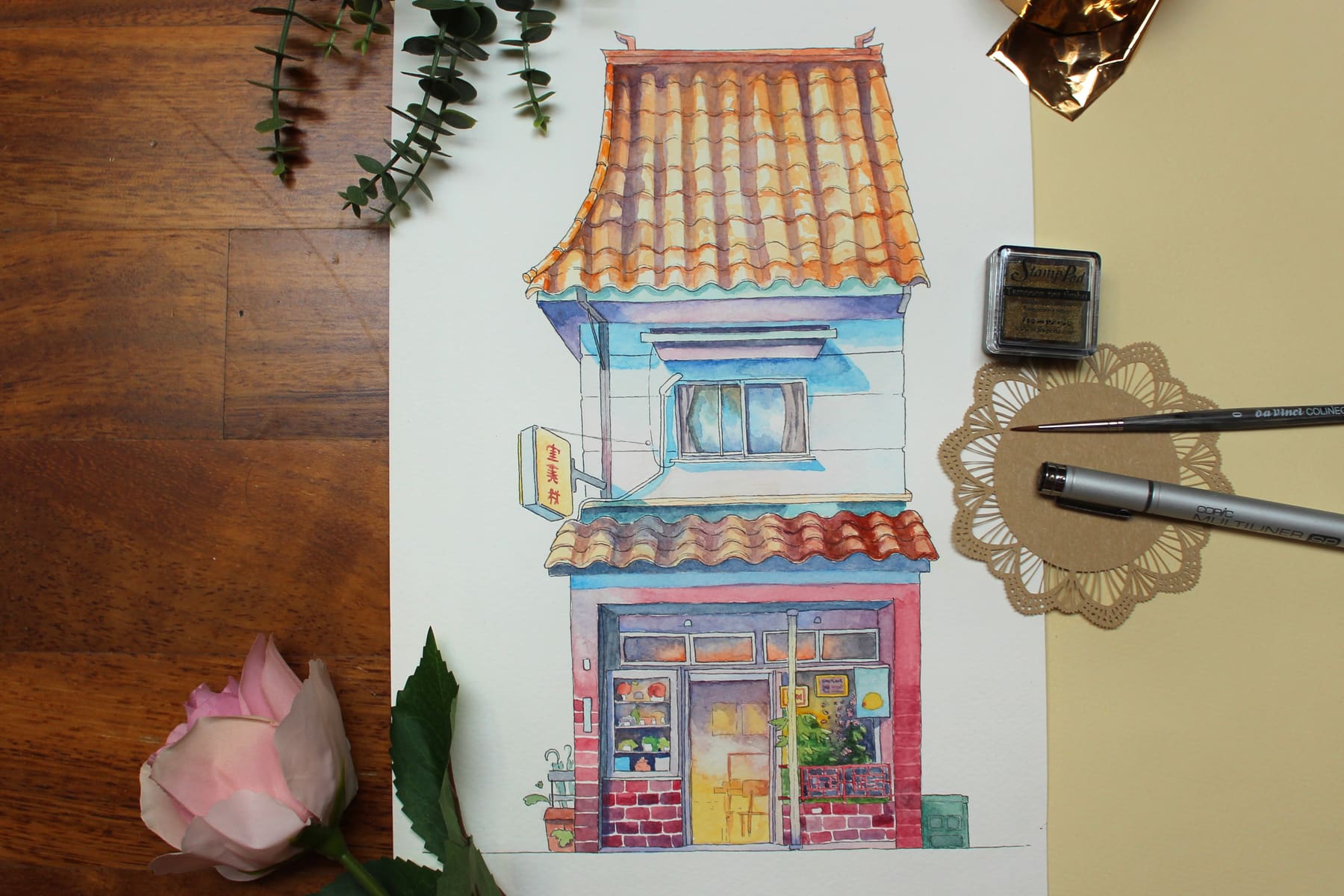 Bottega dall'insegna gialla — l'acquerello intero sul tavolo di legno, con una rosa, un pennello e un tampone per timbri.