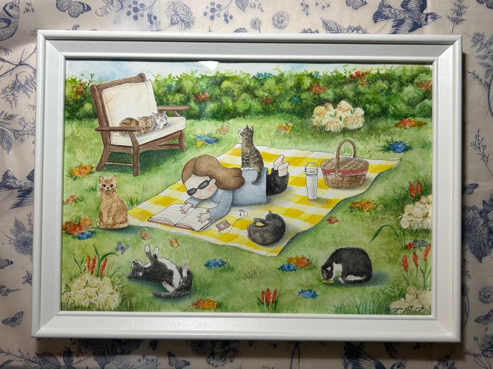 Picnic con i gatti — acquerello su commissione, in cornice bianca: una ragazza legge sdraiata su una coperta a quadri gialli, circondata da sei gatti in giardino.