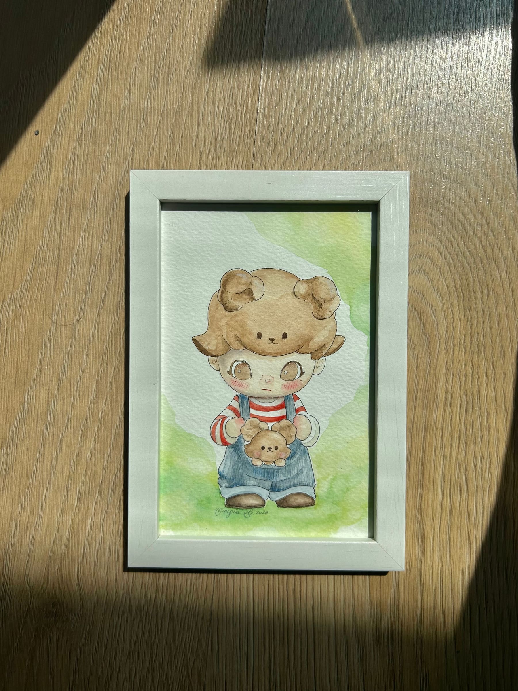 Bear Dad — acquerello originale: un bambino con il cappuccio a orsetto, la maglietta a righe rosse e la salopette di jeans, che stringe un piccolo orsacchiotto su un prato verde.